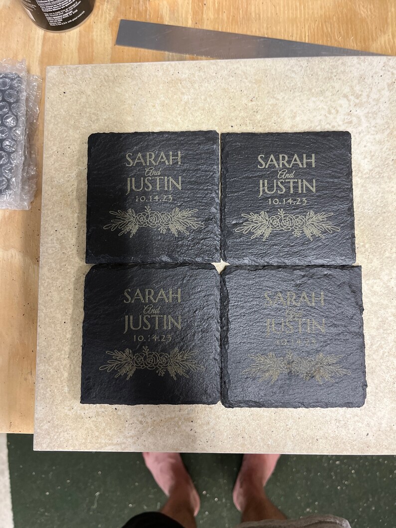 May include: Four black slate coasters with gold lettering. The coasters are engraved with the names "Sarah and Justin" and the date "10.14.23".