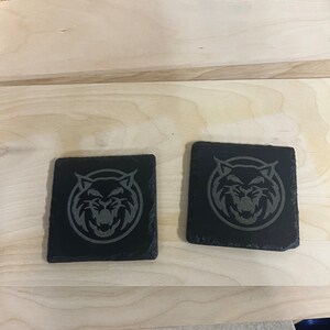 May include: Two black slate coasters featuring a grey engraved image of a wildcat's face.