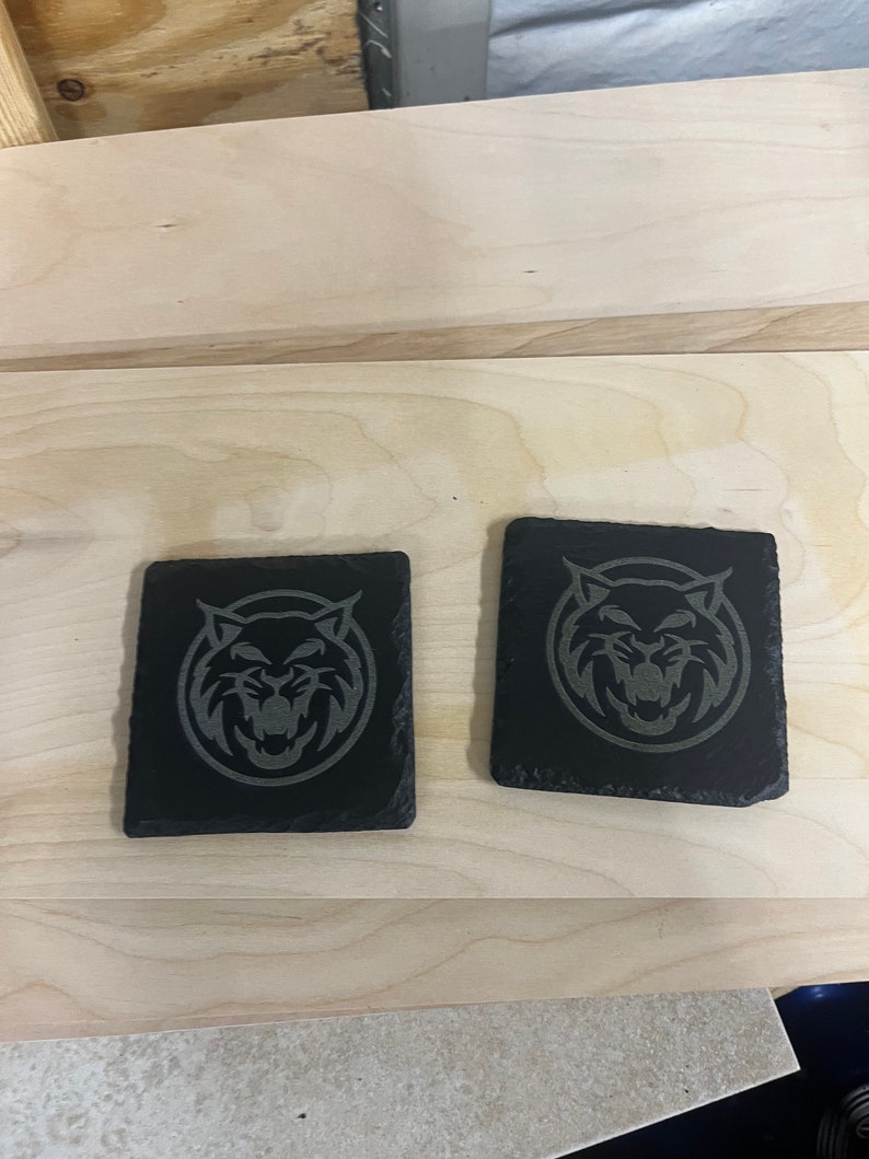 May include: Two black slate coasters featuring a grey engraved image of a wildcat's face.