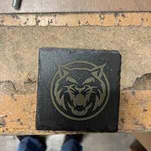 May include: A black slate coaster featuring a gold-coloured graphic of a stylised wildcat head.