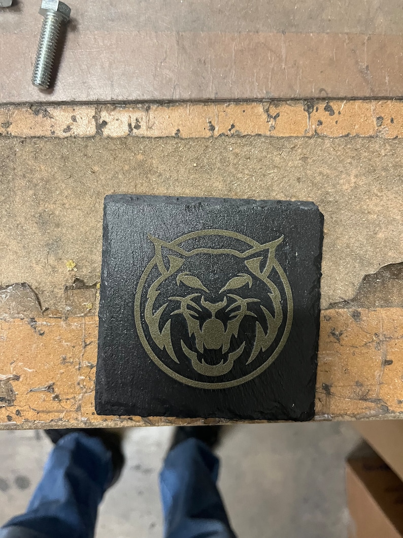 May include: A black slate coaster featuring a gold-coloured graphic of a stylised wildcat head.
