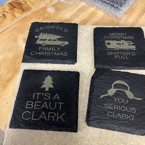 May include: Four black slate coasters with gold lettering. The coasters feature quotes and images from the film 'Christmas Vacation'. The first coaster says 'Griswold Family Christmas' and has a cartoon image of a station wagon with a Christmas tree on top. The second coaster says 'Merry Christmas Shitter's Full'. The third coaster says 'It's a beaut Clark' and has a cartoon image of a Christmas tree. The fourth coaster says 'You serious Clark?' and has a cartoon image of a pair of earmuffs.