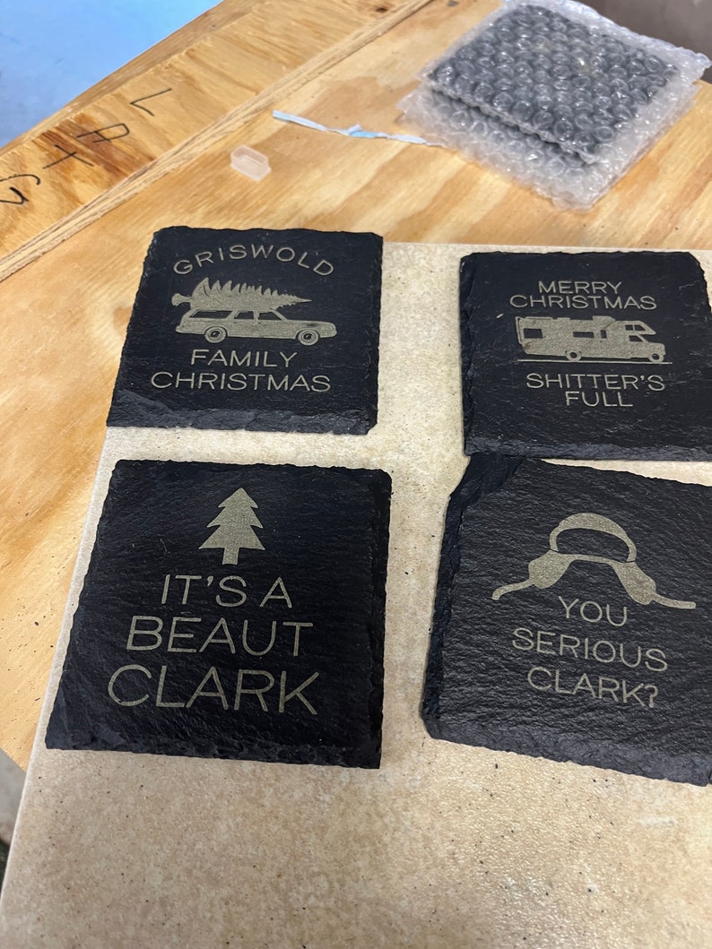 May include: Four black slate coasters with gold lettering. The coasters feature quotes and images from the film 'Christmas Vacation'. The first coaster says 'Griswold Family Christmas' and has a cartoon image of a station wagon with a Christmas tree on top. The second coaster says 'Merry Christmas Shitter's Full'. The third coaster says 'It's a beaut Clark' and has a cartoon image of a Christmas tree. The fourth coaster says 'You serious Clark?' and has a cartoon image of a pair of earmuffs.