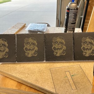 May include: Four black slate coasters featuring a gold design of a woman with a skull face and flowers in her hair.