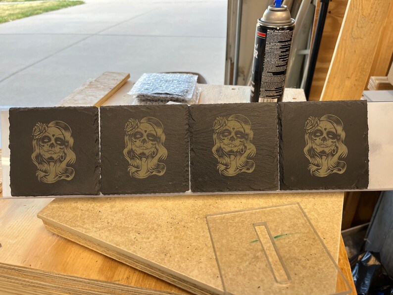 May include: Four black slate coasters featuring a gold design of a woman with a skull face and flowers in her hair.