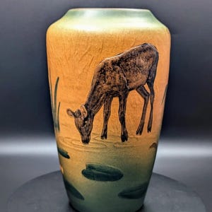 May include: A ceramic vase with a matte finish featuring a gradient from golden yellow to deep green. The surface displays a dark brown illustration of a deer drinking from a pond, surrounded by green lily pads and tall grass blades.