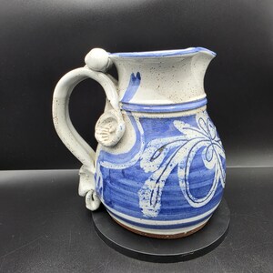 Pode incluir: Um jarro de cerâmica com esmalte esbranquiçado salpicado e padrões florais pintados à mão em azul. O recipiente apresenta uma pega curva com detalhes decorativos em argila, um bico largo e um corpo arredondado com faixas azuis horizontais.