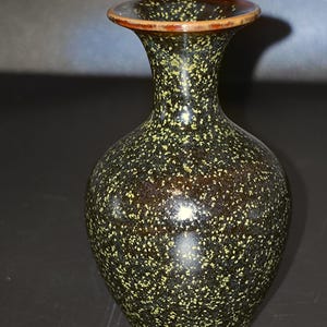 May include: A small ceramic vase with a bulbous body and a flared rim. The surface features a dark brown base color covered in a dense, speckled pattern of light yellow and green dots, with a smooth, glossy finish throughout the entire exterior.