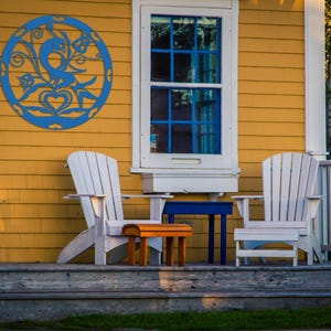 Pode incluir: Duas cadeiras de madeira brancas com uma pequena mesa de madeira e uma mesa de metal azul em uma varanda de madeira com uma casa amarela ao fundo. Uma decora&ccedil;&atilde;o de parede circular de metal azul com um design de p&aacute;ssaro est&aacute; na parede da casa.