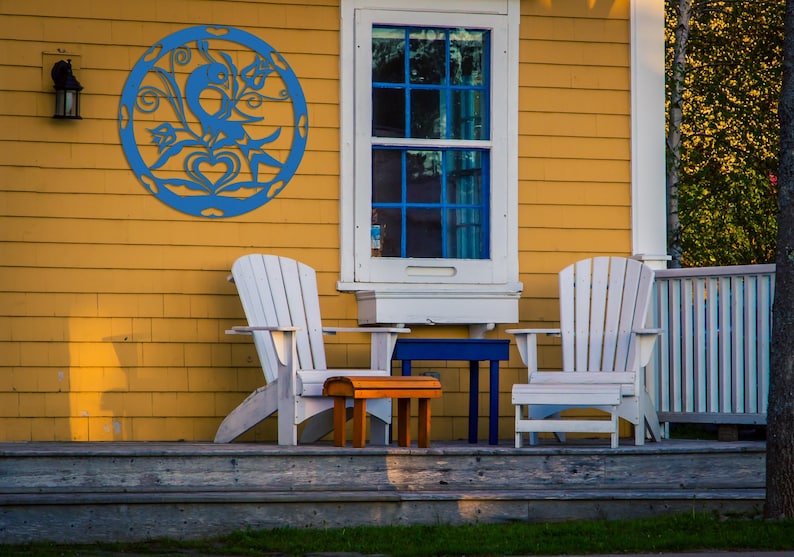 Pode incluir: Duas cadeiras de madeira brancas com uma pequena mesa de madeira e uma mesa de metal azul em uma varanda de madeira com uma casa amarela ao fundo. Uma decora&ccedil;&atilde;o de parede circular de metal azul com um design de p&aacute;ssaro est&aacute; na parede da casa.
