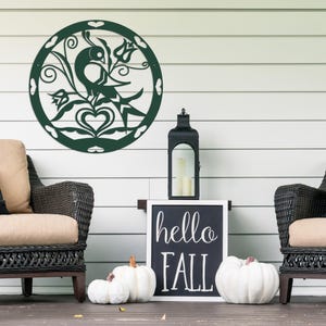 Pode incluir: Uma decora&ccedil;&atilde;o de parede de metal verde com um design de p&aacute;ssaro e flores. A decora&ccedil;&atilde;o est&aacute; pendurada em uma parede branca com duas cadeiras de vime e uma placa que diz "hello fall".