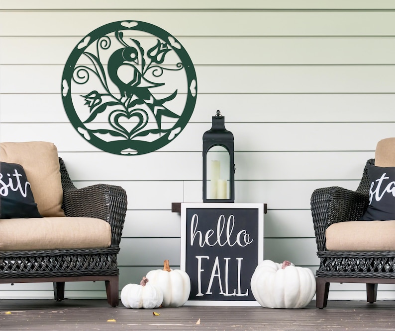 Pode incluir: Uma decora&ccedil;&atilde;o de parede de metal verde com um design de p&aacute;ssaro e flores. A decora&ccedil;&atilde;o est&aacute; pendurada em uma parede branca com duas cadeiras de vime e uma placa que diz "hello fall".