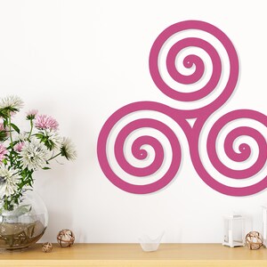 May include: A magenta-colored wall art piece featuring a triple spiral design. The artwork is mounted on a white wall above a wooden table. A clear glass vase with pink and white flowers sits on the table, along with decorative items.