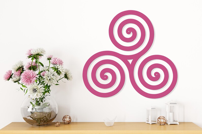 May include: A magenta-colored wall art piece featuring a triple spiral design. The artwork is mounted on a white wall above a wooden table. A clear glass vase with pink and white flowers sits on the table, along with decorative items.