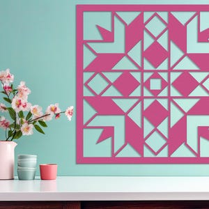 May include: A magenta-colored, geometric metal wall art piece with a quilt-like design. The artwork features cut-out shapes of diamonds, squares, and arrows. A pink pitcher with flowers and small bowls are on a white surface.