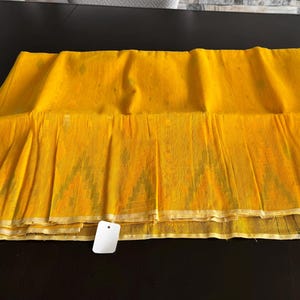 May include: A vibrant yellow fabric textile featuring a subtle, repeating geometric ikat pattern in a lighter shade. The rectangular piece of cloth has a finished hem along the bottom edge and includes a small, blank white rectangular price tag.