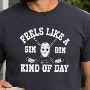 May include: Dark gray t-shirt with the text "FEELS LIKE A SIN BIN KIND OF DAY" in white. The design includes a hockey mask, crossed hockey sticks, and a hockey puck. The shirt is made of a soft material.