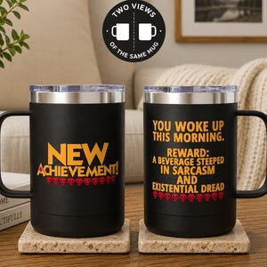 May include: Two black stainless steel mugs with silver rims and lids. The left mug features the yellow text NEW ACHIEVEMENT! above a row of red skulls. The right mug displays the yellow text YOU WOKE UP THIS MORNING. REWARD: A BEVERAGE STEEPED IN SARCASM AND EXISTENTIAL DREAD above a row of red skulls.