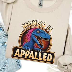 Graphic apparel design featuring a blue feathered raptor with bright pink plumage inside a vintage badge-style emblem. The raptor displays a mischievous expression with sharp teeth and golden eyes, while bold lettering reads “MONGO IS APPALLED.” Inspired by a beloved fantasy book character, this humorous design is perfect for fans of adventure novels, dungeon-crawling stories, and sarcastic monster-themed apparel. Available on a t-shirt, sweatshirt, or hoodie.