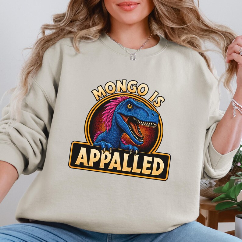 Graphic apparel design featuring a blue feathered raptor with bright pink plumage inside a vintage badge-style emblem. The raptor displays a mischievous expression with sharp teeth and golden eyes, while bold lettering reads “MONGO IS APPALLED.” Inspired by a beloved fantasy book character, this humorous design is perfect for fans of adventure novels, dungeon-crawling stories, and sarcastic monster-themed apparel. Available on a t-shirt, sweatshirt, or hoodie.