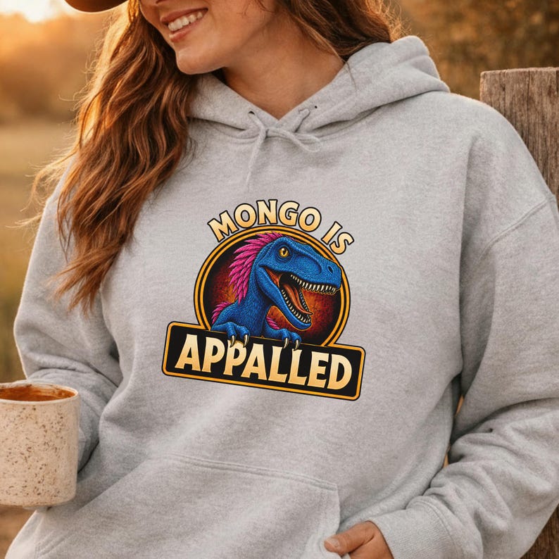 Graphic apparel design featuring a blue feathered raptor with bright pink plumage inside a vintage badge-style emblem. The raptor displays a mischievous expression with sharp teeth and golden eyes, while bold lettering reads “MONGO IS APPALLED.” Inspired by a beloved fantasy book character, this humorous design is perfect for fans of adventure novels, dungeon-crawling stories, and sarcastic monster-themed apparel. Available on a t-shirt, sweatshirt, or hoodie.