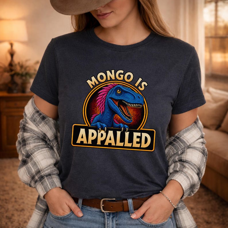 Graphic apparel design featuring a blue feathered raptor with bright pink plumage inside a vintage badge-style emblem. The raptor displays a mischievous expression with sharp teeth and golden eyes, while bold lettering reads “MONGO IS APPALLED.” Inspired by a beloved fantasy book character, this humorous design is perfect for fans of adventure novels, dungeon-crawling stories, and sarcastic monster-themed apparel. Available on a t-shirt, sweatshirt, or hoodie.