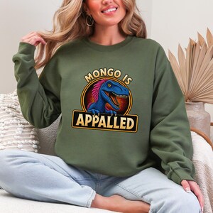 Graphic apparel design featuring a blue feathered raptor with bright pink plumage inside a vintage badge-style emblem. The raptor displays a mischievous expression with sharp teeth and golden eyes, while bold lettering reads “MONGO IS APPALLED.” Inspired by a beloved fantasy book character, this humorous design is perfect for fans of adventure novels, dungeon-crawling stories, and sarcastic monster-themed apparel. Available on a t-shirt, sweatshirt, or hoodie.