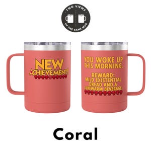 May include: A coral-coloured stainless steel mug with a handle and lid. One side displays the text NEW ACHIEVEMENT! with a row of red skulls. The other side reads YOU WOKE UP THIS MORNING. REWARD: MILD EXISTENTIAL DREAD AND A LUKEWARM BEVERAGE.