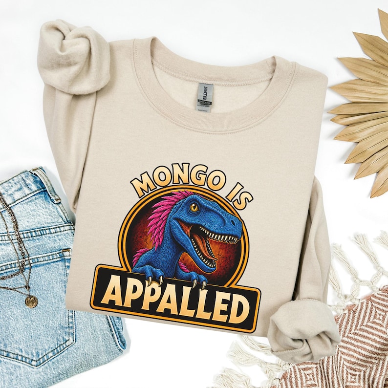 Graphic apparel design featuring a blue feathered raptor with bright pink plumage inside a vintage badge-style emblem. The raptor displays a mischievous expression with sharp teeth and golden eyes, while bold lettering reads “MONGO IS APPALLED.” Inspired by a beloved fantasy book character, this humorous design is perfect for fans of adventure novels, dungeon-crawling stories, and sarcastic monster-themed apparel. Available on a t-shirt, sweatshirt, or hoodie.