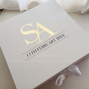 May include: A cream-coloured gift box with gold lettering displaying the initials "SA" and the date "11TH FEBRUARY 2024". A white ribbon is tied on the side. Suitable for a special event.