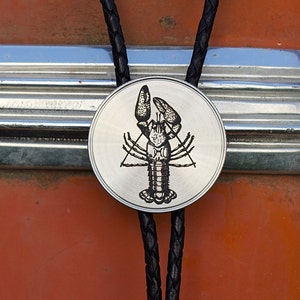 May include: A silver-colored bolo tie with a detailed black lobster design on a round pendant. The pendant is attached to a black braided leather cord, creating a unique accessory.