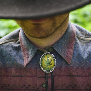 May include: Oval bolo tie with a silver-colored frame and a green cannabis leaf design on a yellow background. The tie features a black leather cord and is set against a patterned button-down shirt in shades of burgundy, blue, and green.