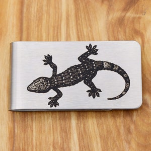 May include: A brushed metal money clip featuring a detailed black and white illustration of a gecko. The gecko design is centered on the clip, with a textured appearance. The clip is rectangular with rounded corners.
