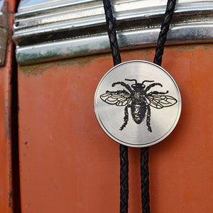 May include: A silver-colored bolo tie with a black braided cord. The circular pendant features a detailed black illustration of a bee. The background is a weathered orange surface.
