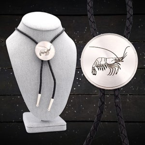 May include: A silver bolo tie featuring a detailed shrimp design. The round pendant is attached to a black braided leather cord with silver tips. The bolo tie is displayed on a gray mannequin.