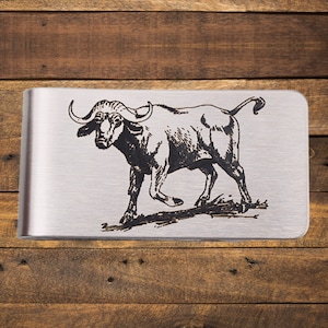 May include: A silver-toned money clip with a detailed black engraving of a buffalo. The buffalo is depicted in a running pose, with its horns and muscular build clearly visible. The clip has a brushed metal finish.