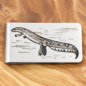 Op de afbeelding: Een geldclip van geborsteld metaal met een zwart-wit illustratie van een salamander. De salamander wordt zwemmend afgebeeld, met uitgestrekte poten en een gebogen lichaam. De clip is rechthoekig met afgeronde hoeken.