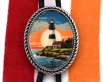 May include: A silver bolo tie with a black leather cord and a round glass pendant featuring a lighthouse scene with a sunset and ocean waves.
