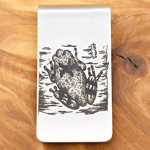 May include: A silver-colored money clip with a detailed black illustration of a frog. The frog is depicted in a pond setting, with water and plant details. The clip is rectangular with rounded edges.