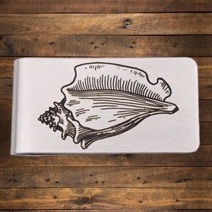 May include: A silver money clip with a black conch shell illustration. The shell design is etched onto the clip's surface, showing the natural curves and textures of the shell. The clip is made of metal.
