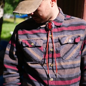 Puede incluir: Un bolo tie rojo y negro con una concha de plata. La concha tiene un patrón rojo, negro y blanco. El bolo tie lo lleva una persona que lleva una camisa de cuadros rojos y negros.