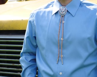 Könnte beinhalten: Ein hellblaues Hemd mit Knöpfen und einer Bolo-Krawatte mit einem silbernen und blauen Adler-Design. Die Bolo-Krawatte besteht aus einem braunen Lederband mit silbernen Enden.