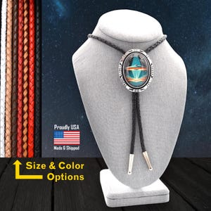 Könnte beinhalten: Ein schwarzes Bolo-Band mit einem ovalen Silberanhänger, der ein farbenfrohes Bild eines UFOs zeigt. Der Anhänger hat einen silbernen Rand mit einem indianischen Design. Das Bolo-Band wird auf einer grauen Schaufensterpuppe präsentiert.