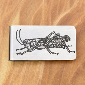 May include: A silver money clip featuring a detailed black engraving of a grasshopper. The insect is shown in profile, with its legs, antennae, and body segments clearly visible. The clip has rounded corners.
