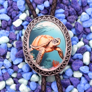 May include: A silver bolo tie with a round, detailed, silver frame featuring a colorful image of a sea turtle swimming in blue water with coral and seaweed. The bolo tie is on a bed of blue and white stones.