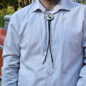 Puede incluir: Una corbata bolo plateada con un colgante circular con un diseño detallado. La corbata bolo tiene un cordón negro con puntas plateadas y se usa con una camisa abotonada azul claro estampada.