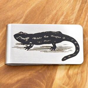 Op de afbeelding: Een geldclip van geborsteld metaal met een gedetailleerde zwarte illustratie van een salamander. De salamander is in profiel afgebeeld, met ingewikkelde details op zijn lichaam en staart. De clip is rechthoekig en heeft een eenvoudig ontwerp.