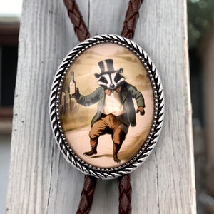May include: A silver bolo tie with a round, glass pendant featuring a cartoon badger wearing a top hat and holding a bottle of alcohol. The badger is standing on a path in a field.