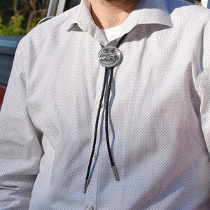 Pode incluir: Uma camisa branca abotoada com uma gravata bolo. A gravata bolo tem um pendente circular prateado com um desenho. O cordão da gravata é preto e trançado.