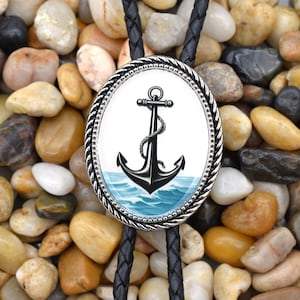 Könnte beinhalten: Ein schwarzer Bolo-Krawattenhalter mit einem silberfarbenen Anker-Anhänger. Der Anker ist vor einem blauen Wellenhintergrund platziert und von einem seilartigen Rand umgeben.